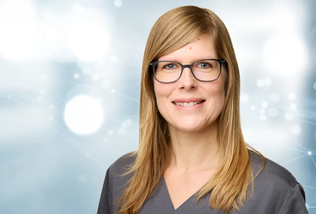 Porträt von Frau mit schulterlangen dunkelblonden Haaren und schwarzer Brille. Vor blauem Hintergrund. Sie lächelt.